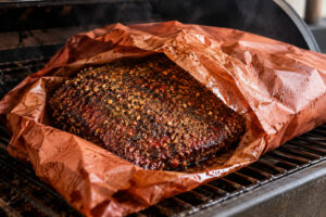 Pastrami mit dunkler Gewürzkruste in geöffnetem Butcher Paper auf dem Rost eines Smokers